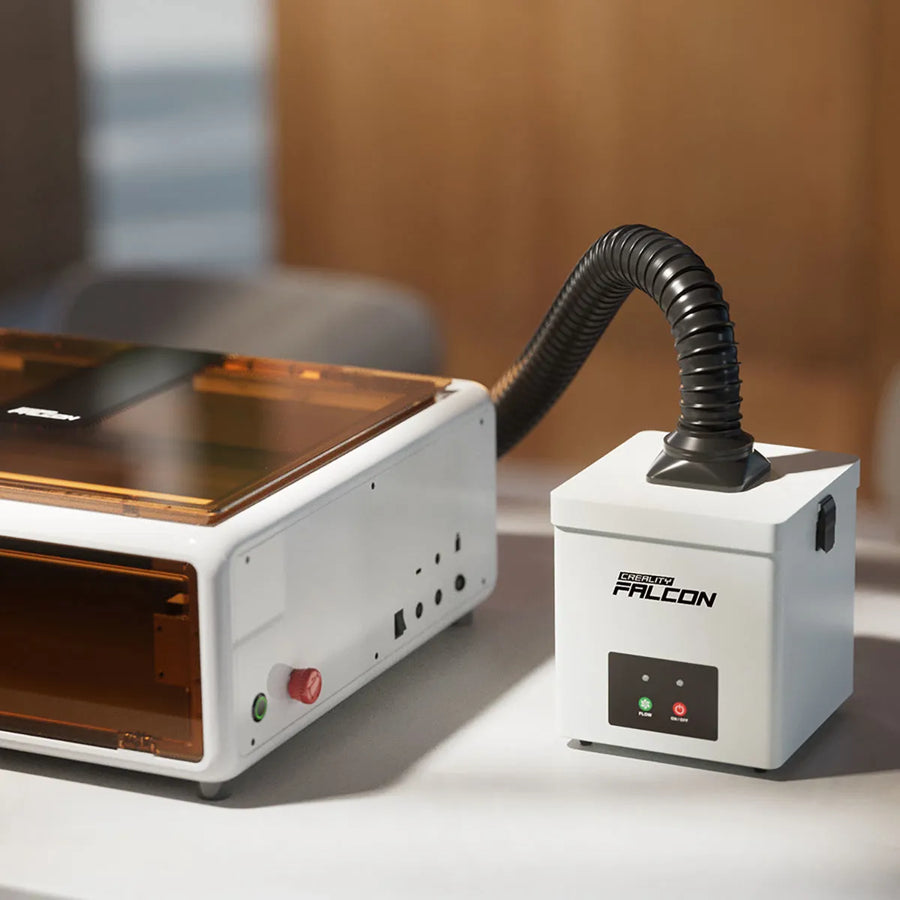 Creality Falcon smoke purifier with a black flexible hose connected, placed beside a white enclosed laser engraver on a desk